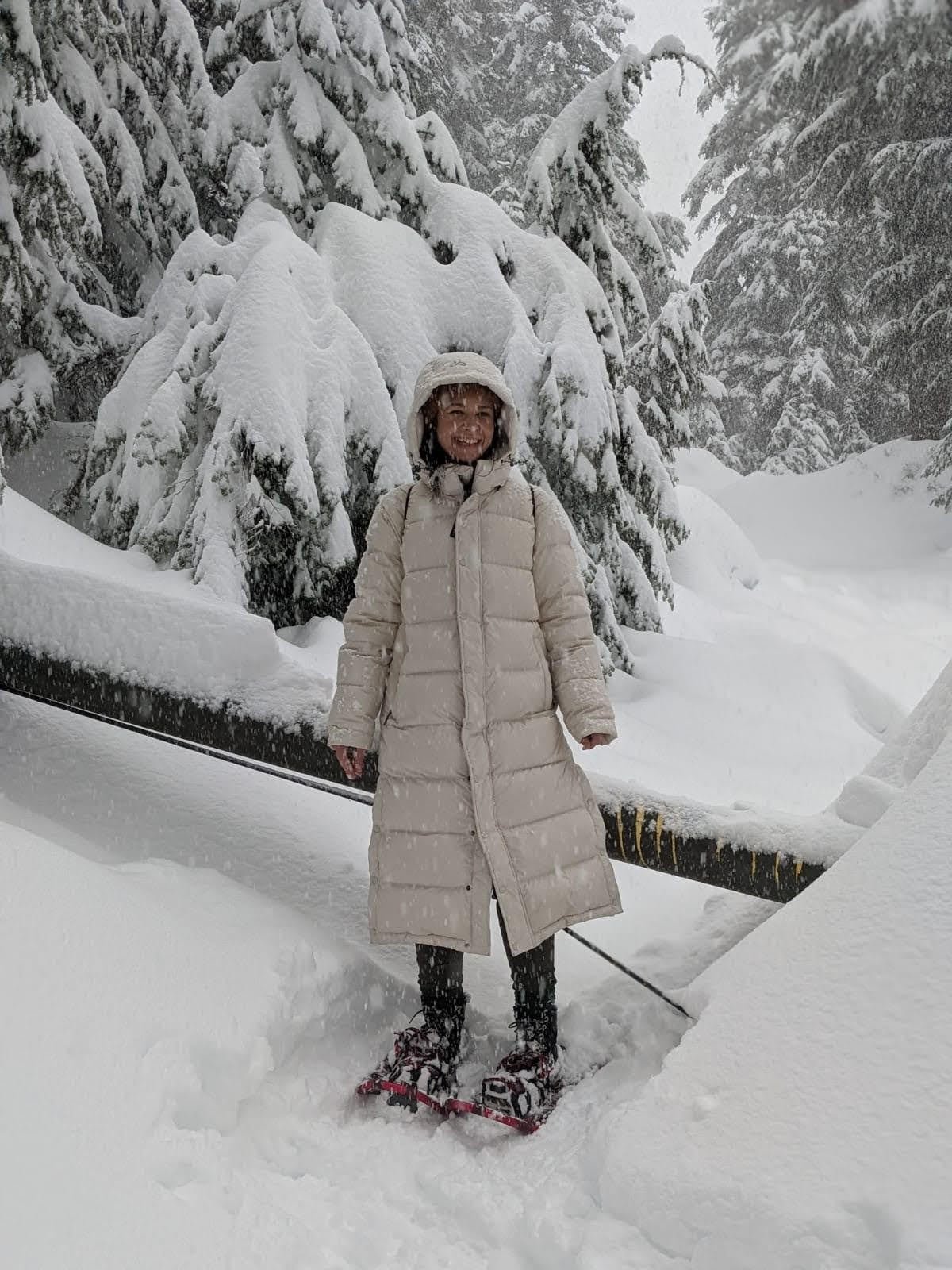 default-16 A woman in a white poofy jacket and snowshoes smiles as she poses on a snow-covered trail through tall pine trees.