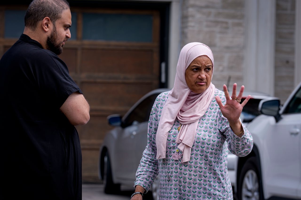 A woman standing outside a residence, gesturing with her hand next to a man, with two cars in the background in the driveway.